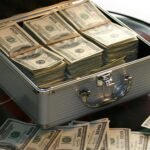 Open briefcase filled with stacks of hundred dollar bills on a glass table, representing wealth.